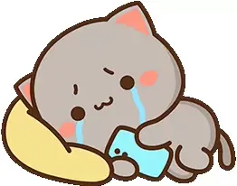 A gray mochi cat, Goma, is crying while looking at a phone. The cat is leaning on a yellow pillow and looks sad.